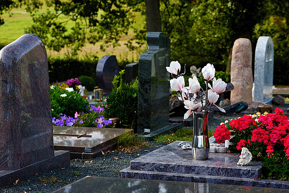Mehrere Grabsteine auf einem Friedhof auf einer Grabplatte im Vordergrund steht eine chromfarbene hohe Vase mit Magnolienblüten darin