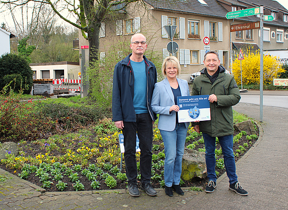 Drei Personen die ein Schild halten stehen vor einem Blumenbeet mit neu gepflanzten Vergissmeinnicht