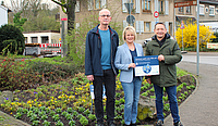 Drei Personen die ein Schild halten stehen vor einem Blumenbeet mit neu gepflanzten Vergissmeinnicht