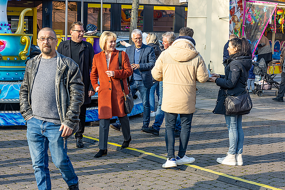 Fünf Personen bewegen sich über die Kirmes, lächelnd, sie unterhalten sich miteinander und mit einem an sie herantretenden jungen Paar.