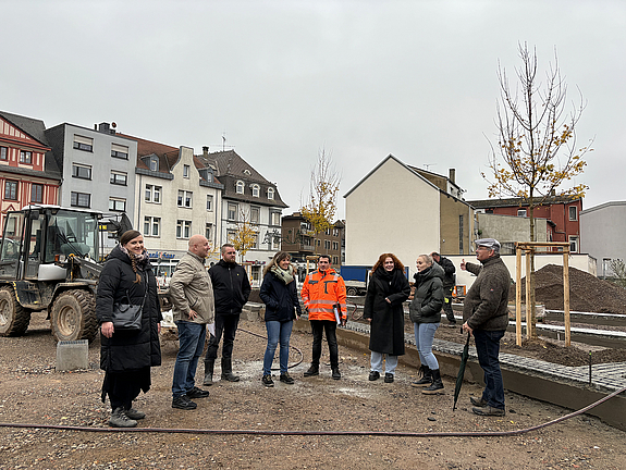 Sieben Personen stehen in einem Halbkreis in einem Baufeld, hinter ihnen steht ein Bagger, man sieht drei neu gepflanzten Bäume, die sich noch in einer Stützkonstuktion aus Holz befinden.