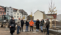 Sieben Personen stehen in einem Halbkreis in einem Baufeld, hinter ihnen steht ein Bagger, man sieht drei neu gepflanzten Bäume, die sich noch in einer Stützkonstuktion aus Holz befinden.