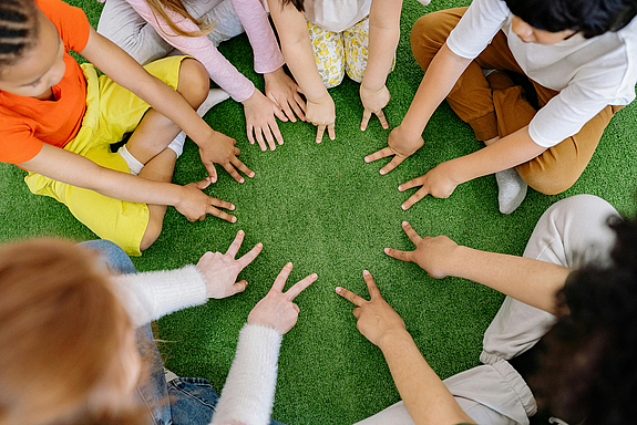 Kinder bilden sitzend einen Kreis und stützen ihre gespreizten Finger auf eine Grasmatte