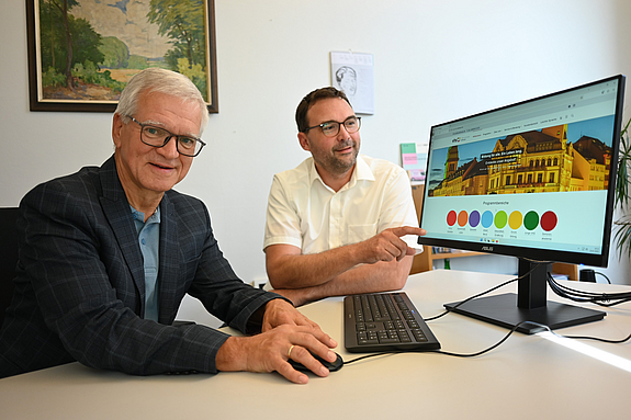 Oberbürgermeister und VHS-Direktor sitzen zusammen vor einem PC-Bildschirm auf dem die neue Webseite der VHS zu sehen ist.
