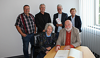 Der Künstler und seine Frau sitzen an einem Tisch mit dem aufgeschlagenen Goldenen Buch vor sich im Büro des Oberbürgermeisters. Die vier anderen Personen stehen hinter ihnen. 