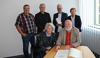 Der Künstler und seine Frau sitzen an einem Tisch mit dem aufgeschlagenen Goldenen Buch vor sich im Büro des Oberbürgermeisters. Die vier anderen Personen stehen hinter ihnen. 