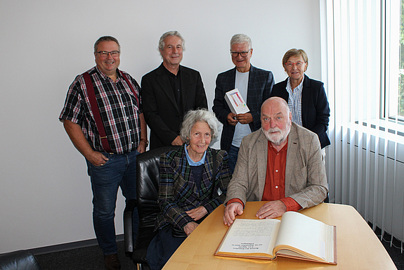 Der Künstler und seine Frau sitzen an einem Tisch mit dem aufgeschlagenen Goldenen Buch vor sich im Büro des Oberbürgermeisters. Die vier anderen Personen stehen hinter ihnen. 