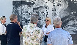 Vier Personen stehen vor einem großen Mural mit dem Rücken zum Betrachter, eine fünfte Person referiert