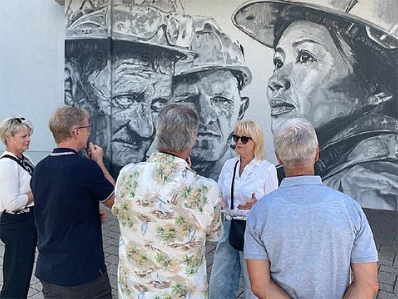 Vier Personen stehen vor einem großen Mural mit dem Rücken zum Betrachter, eine fünfte Person referiert