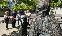 Eine Skulptur im Vordergrund, im Hintergrund stehen vier Personen die einer fünften Person zugewandt zuhören