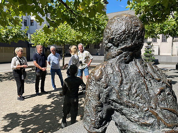Eine Skulptur im Vordergrund, im Hintergrund stehen vier Personen die einer fünften Person zugewandt zuhören
