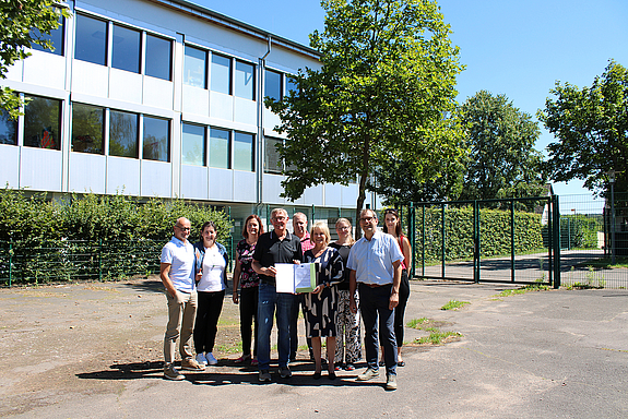 Auf dem asphaltierten Bereich der Grundschule Lauterbach stehen neun Personen, die Oberbürgermeisterin und der LAG Warndt-Saargau Vorsitzende halten den Förderbescheid in Händen